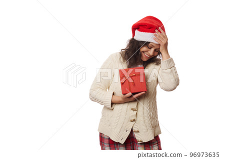 Waist-up portrait of a beautiful multi-ethnic positive woman, wearing Santa hat and warm knitted sweater, rejoicing at happy Christmas present, isolated over white background. Copy advertising space 96973635