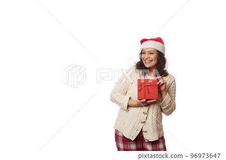 Waist-up portrait on white background of a multi-ethnic positive pretty woman wearing Santa hat, cheerfully smiling looking aside at copy ad space, posing with gift box with Christmas happy present 96973647