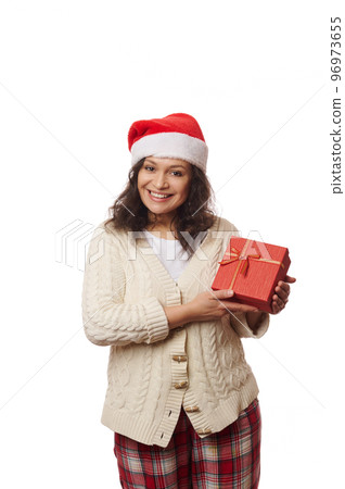 Waist-up portrait on white background of a positive cheerful woman in Santa hat, beige warm knitted sweater and checkered trousers, smiling looking at camera, posing with happy present for Christmas 96973655