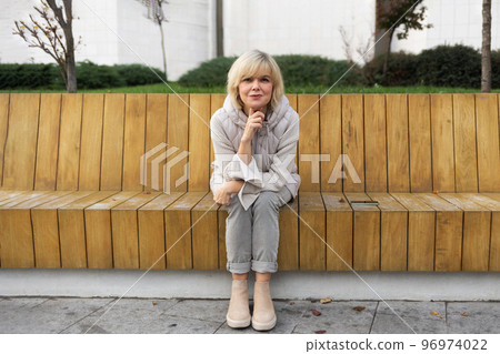 Attractive Caucasian adult smiling blonde woman in jacket posing sitting on bench in city park. Concept of carefree and psychology Attractive Caucasian adult smiling blonde woman in jacket posing sitting on bench in city park. Concept of carefree and psychology 96974022