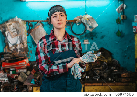 Portrait of a young female mechanic in uniform, wearing gloves. Working room in the background Portrait of a young female mechanic in uniform, wearing gloves. Working room in the background 96974599