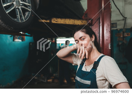 Gender equality. A young brunette in uniform stands near the Elevator with a car and wipes her nose with her hand. In the background is an auto repair shop 96974600