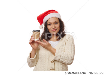 Isolated on white background, attractive positive multi-ethnic brunette woman, wearing Santa hat, holding takeaway hot tea or coffee in recyclable disposable eco paper cup. Food and drink consumerism. 96976172