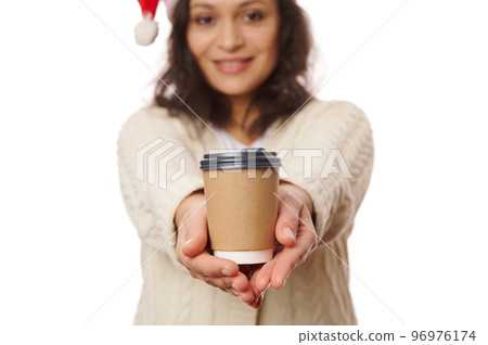 Selective focus on a takeaway hot drink in disposable cardboard cup in the hands of a blurred woman in Santa hat, smiling on camera, isolated over white background. Beverage to go. Mockup. Copy space. Selective focus on a takeaway hot drink in disposable cardboard cup in the hands of a blurred woman in Santa hat, smiling on camera, isolated over white background. Beverage to go. Mockup. Copy space. 96976174