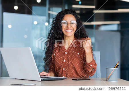 Portrait successful Hispanic business woman office worker with curly hair smiling and looking at camera working at table with laptop holding hand up gesture success and triumph celebrating victory 96976211