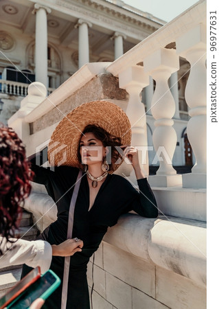 Fashion photo of a beautiful model in an elegant hat and black dress posing against the backdrop of a building with columns. Fashion photo of a beautiful model in an elegant hat and black dress posing against the backdrop of a building with columns. 96977631
