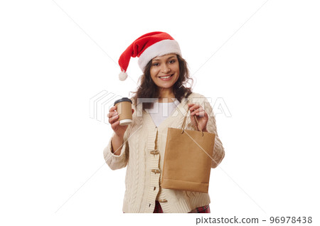 Multi-ethnic charming woman in Santa hat, unpacking an eco paper bag with takeaway food and drink, cutely smiling on camera, isolated over white background. Beverage to go. Delivery at Christmastime 96978438