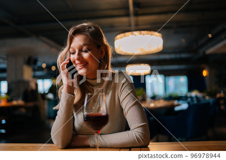Portrait of cheerful blonde female talking on mobile phone sitting at table with red wine in modern restaurant with dark interior. Attractive happy young woman having conversation on smartphone in bar Portrait of cheerful blonde female talking on mobile phone sitting at table with red wine in modern restaurant with dark interior. Attractive happy young woman having conversation on smartphone in bar 96978944