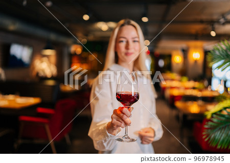 Selective focus portrait of blonde woman holding glass of red wine standing in restaurant with luxury interior, looking at camera. Front view of cute female posing at cafe, blurred background 96978945