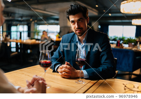 View from back of unrecognizable young woman to smiling elegant man in suit sitting at table with glasses of red wine having festive dinner at luxury restaurant. Happy loving couple enjoying talking. 96978946