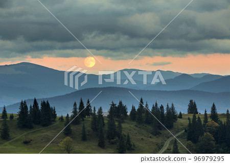 Covered with mist mountains at moonrise, big moon on bright orange sky over tall fir-trees. Covered with mist mountains at moonrise, big moon on bright orange sky over tall fir-trees. 96979295