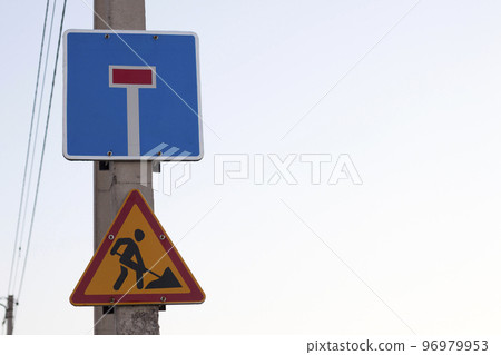 Bright yellow triangular warning sign; Attention, work ahead, and Dead-end street on square blue shield on concrete pillar on clear blue sky copy space background. Repair, road construction concept. 96979953