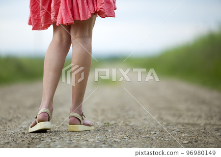 Back view of young woman in red dress legs walking by ground road on summer day on blurred sunny background. Back view of young woman in red dress legs walking by ground road on summer day on blurred sunny background. 96980149