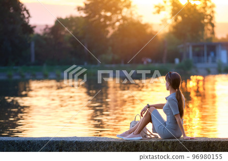 Back view of lonely young woman sitting alone on lake shore enjoying warm evening. Wellbeing and relaxing in nature concept. Back view of lonely young woman sitting alone on lake shore enjoying warm evening. Wellbeing and relaxing in nature concept. 96980155