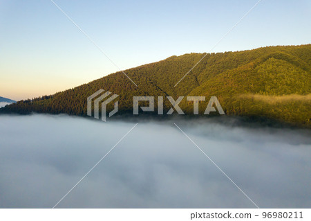 Aerial view of vibrant sunrise over white dense fog with distant dark silhouettes of mountain hills on horizon. 96980211
