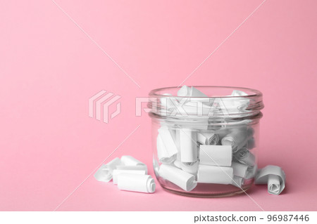Glass jar full of rolled paper sheets on pink background, space for text 96987446
