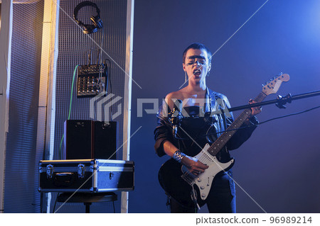 Stylish guitarist playing alternative guitar and performing heavy metal concert, using microphone to sing cool song and instrument. Artist feeling ecstatic about live rock performance, crazy make up. Stylish guitarist playing alternative guitar and performing heavy metal concert, using microphone to sing cool song and instrument. Artist feeling ecstatic about live rock performance, crazy make up. 96989214
