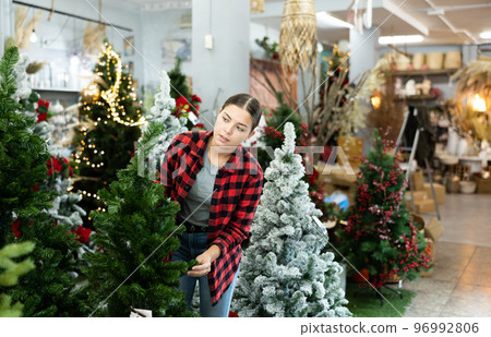 Focused young female choosing an artificial Christmas tree in decorated hall at the December 96992806