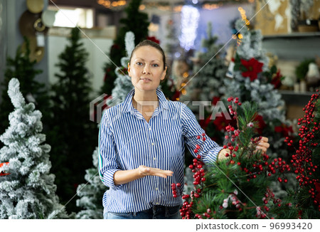 Smiling casual woman checking the material of a Christmas tree with viburnum before New Year in a shopping mall Smiling casual woman checking the material of a Christmas tree with viburnum before New Year in a shopping mall 96993420