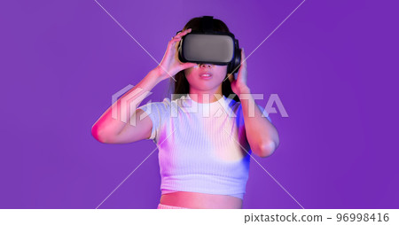 Young asian woman in white t-shirt wearing vr headset looking playing touching purple screen color background. Metaverse concept. Young asian woman in white t-shirt wearing vr headset looking playing touching purple screen color background. Metaverse concept. 96998416