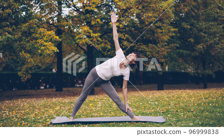 Side view of slender girl is sports clothing exercising outdoors doing yoga in park alone on warm autumn day. Nature, recreation and modern youth concept. Side view of slender girl is sports clothing exercising outdoors doing yoga in park alone on warm autumn day. Nature, recreation and modern youth concept. 96999838