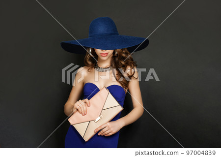 Young Smiling Woman in Blue Dress and wide blue broad brim hat. Fashion Model with Wavy Brown Hair Young Smiling Woman in Blue Dress and wide blue broad brim hat. Fashion Model with Wavy Brown Hair 97004839