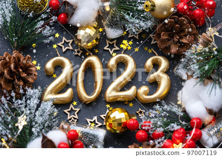 The golden figures 2023 made of candles on a black stone slate background are decorated with a festive decor of stars, sequins, fir branches, balls and garlands. Greeting card, happy New Year. 97007119
