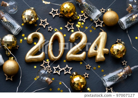 The golden figures 2024 made of candles on a black stone slate background are decorated with a festive decor of stars, sequins, fir branches, balls and garlands. Greeting card, happy New Year. 97007127