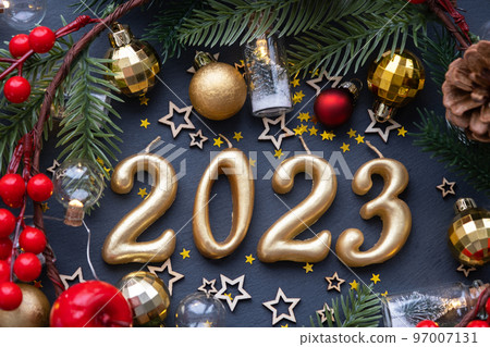 The golden figures 2023 made of candles on a black stone slate background are decorated with a festive decor of stars, sequins, fir branches, balls and garlands. Greeting card, happy New Year. The golden figures 2023 made of candles on a black stone slate background are decorated with a festive decor of stars, sequins, fir branches, balls and garlands. Greeting card, happy New Year. 97007131
