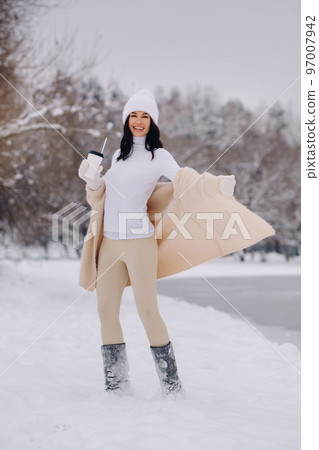 A beautiful girl in a beige cardigan and a white hat with a glass of tea enjoys a snowy embankment by the lake A beautiful girl in a beige cardigan and a white hat with a glass of tea enjoys a snowy embankment by the lake 97007942