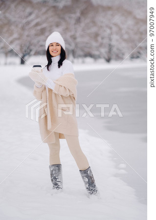 A beautiful girl in a beige cardigan and a white hat with a glass of tea enjoys a snowy embankment by the lake 97007949
