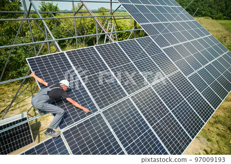 Worker installing solar panels in field at sunny day. European man wearing helmet and uniform. Photo-voltaic collection of modules as a PV panel. Idea of environment safety. Green energy. 97009193