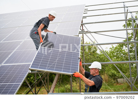Male worker in safety construction helmet giving solar module to colleague. Two men in workwear building photovoltaic solar panel system. Concept of sustainable energy and solar panel installation. 97009202