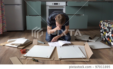 Young man reading instruction while assembling furniture for new apartment on the floor Young man reading instruction while assembling furniture for new apartment on the floor 97011895