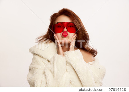 Portrait of young beautiful red-haired woman posing in fur coat and red trendy glasses isolated over white background. Kisses 97014528