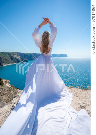 Blonde with long hair on a sunny seashore in a white flowing dress, rear view, silk fabric waving in the wind. Against the backdrop of the blue sky and mountains on the seashore. 97014864