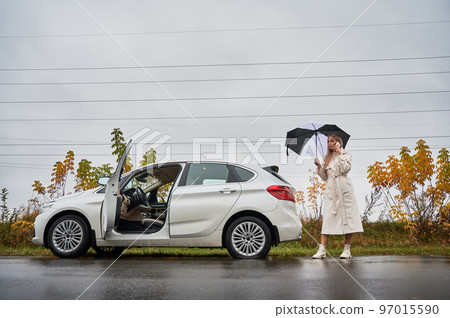 Worried woman standing on road near her white car with punctured car tire. Female driver calling car service for help, holding umbrella. Worried woman standing on road near her white car with punctured car tire. Female driver calling car service for help, holding umbrella. 97015590