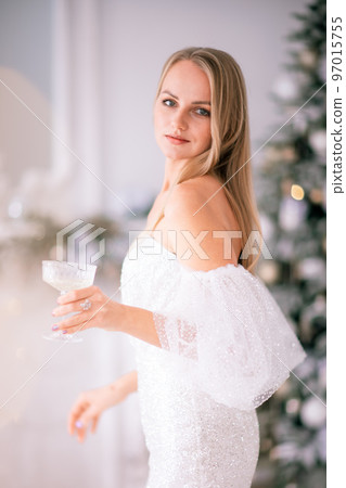 The blonde in the Christmas room. A beautiful blonde woman in a shiny light short dress with a train stands in a beautiful bright room decorated with a festive interior with a Christmas tree. The blonde in the Christmas room. A beautiful blonde woman in a shiny light short dress with a train stands in a beautiful bright room decorated with a festive interior with a Christmas tree. 97015755