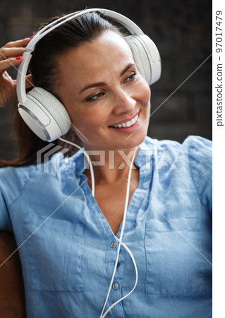 Leisure time concept. Happy beautiful woman listening to the music using headphones sitting on a couch indoors. Female spending her free day and relaxing at home alone. 97017479