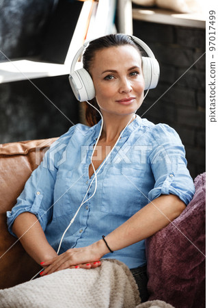 Leisure time concept. Happy beautiful woman listening to the music using headphones sitting on a couch indoors. Female spending her free day and relaxing at home alone. 97017499