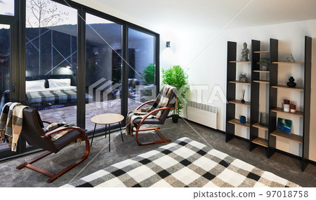 Stylish bedroom with comfortable furniture and irregular panoramic window at twilight. Cozy modern furnished cottage room with reflecting in large curtainless window in evening. Concept of interior. 97018758