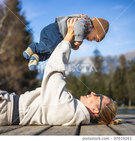 Happy family. Young mother playing with her baby boy infant oudoors on sunny autumn day. Portrait of mom and little son on wooden platform by lake. Positive human emotions, feelings, joy Happy family. Young mother playing with her baby boy infant oudoors on sunny autumn day. Portrait of mom and little son on wooden platform by lake. Positive human emotions, feelings, joy 97019261