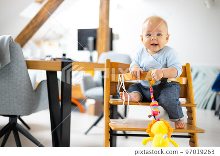 Happy infant sitting and playing with his toy in traditional scandinavian designer wooden high chair in modern bright atic home. Cute baby 97019263