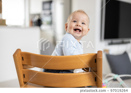 Happy infant sitting in traditional scandinavian designer wooden high chair and laughing out loud in modern bright home. Cute baby smile 97019264