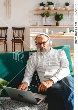 Handsome old man dressed in hoodie and eyeglasses is using laptop and smiling while listen music sitting on couch at home 97022363