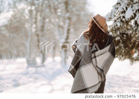 Smiling woman enjoying winter moments in snowy park. Young woman wearing hat, plaid scarf and coat. 97025476