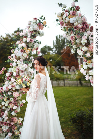 Beautiful fiancee in white wedding dress with bouquet stand near the wedding arch of flowers. 97027019