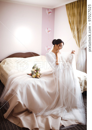 Morning of beautiful bride near the wedding dress. Bride in a white robe holds and admires her wedding dress. 97027025