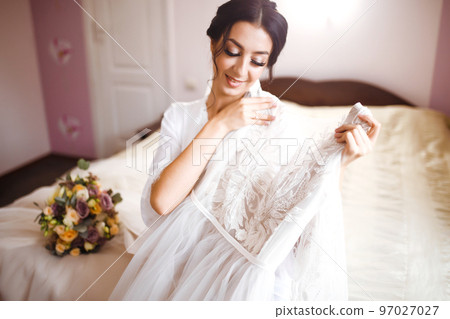 Morning of beautiful bride near the wedding dress. Bride in a white robe holds and admires her wedding dress. 97027027