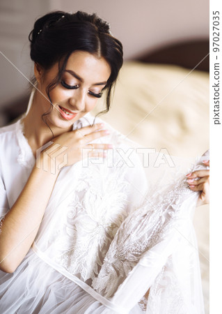 Morning of beautiful bride near the wedding dress. Bride in a white robe holds and admires her wedding dress. 97027035